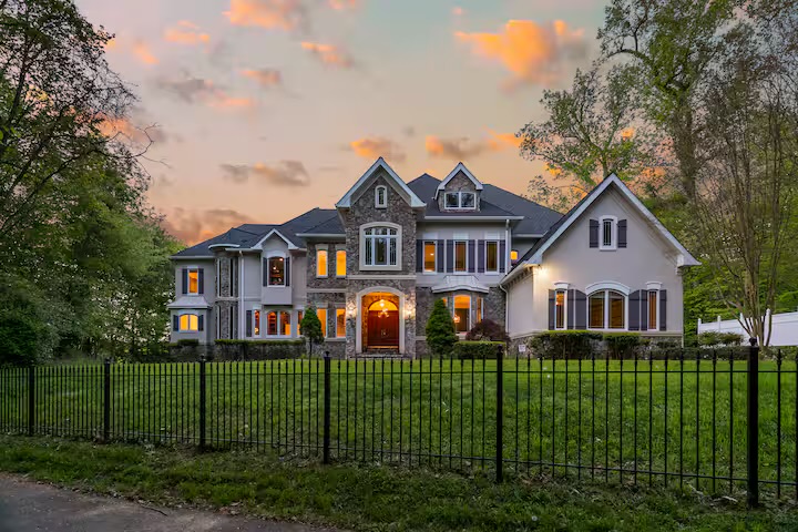 The waterfront estate at dusk, warmly lit windows behind an iron fence